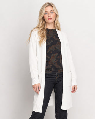 A blonde woman modeling a long white open-front cardigan over a dark patterned top and black jeans, photographed against a plain light gray studio background.