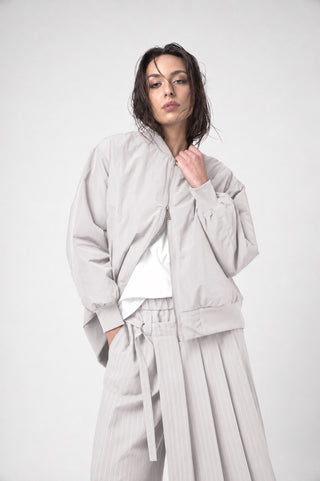 A woman modeling a light beige/taupe two-piece outfit consisting of a zip-up bomber jacket and matching pleated trousers, styled over a white t-shirt, against a plain light gray background.