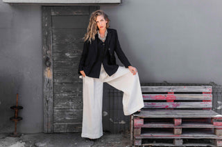 Woman in a black blazer and white pants standing next to a wooden pallet against a gray wall.