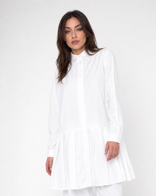 A woman modeling a white long-sleeve collared shirt dress with a pleated hem against a plain white studio background.