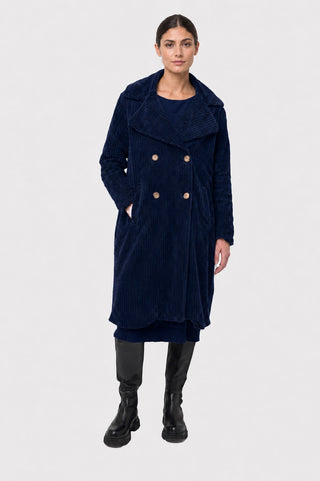 A model stands against a light gray studio background wearing a long navy blue corduroy double-breasted coat with large notched lapels and gold-tone buttons, paired with black boots.