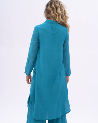 A woman modeling a blue two-piece outfit consisting of a long-sleeve button-front tunic and matching wide-leg pants, photographed against a plain light gray studio background.