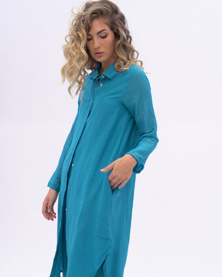 A woman modeling a blue two-piece outfit consisting of a long-sleeve button-front tunic and matching wide-leg pants, photographed against a plain light gray studio background.