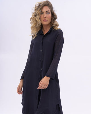 A woman modeling a dark blue two-piece outfit consisting of a long-sleeve button-front tunic and matching wide-leg pants, photographed against a plain light gray studio background.
