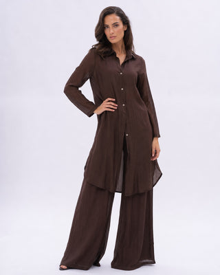 A woman modeling a dark brown two-piece outfit consisting of a long-sleeve button-front tunic and matching wide-leg pants, photographed against a plain light gray studio background.