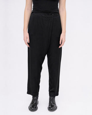 A model wearing black high-waisted, relaxed-fit trousers with a button detail at the waistband, paired with a black top and black leather shoes, shot against a plain white studio background.