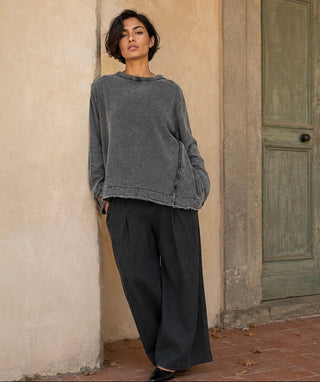 A woman stands outdoors against a beige stucco wall next to a weathered green door, wearing a loose charcoal gray long-sleeve top and dark charcoal wide-leg pants.