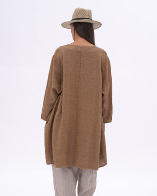 A woman modeling a loose brown long-sleeve tunic top with a V-neck and side pockets, paired with grey pants and a beige straw hat, against a plain white studio background.