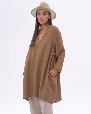 A woman modeling a loose brown long-sleeve tunic top with a V-neck and side pockets, paired with white pants and a beige straw hat, against a plain white studio background.