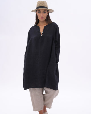 A woman modeling a loose navy long-sleeve tunic top with a V-neck and side pockets, paired with grey pants and a beige straw hat, against a plain white studio background.