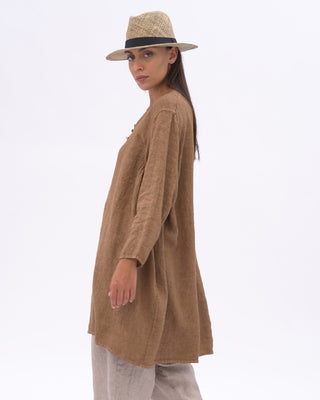 A woman modeling a loose brown long-sleeve tunic top with a V-neck and side pockets, paired with grey pants and a beige straw hat, against a plain white studio background.