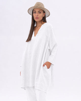 A woman modeling a loose white long-sleeve tunic top with a V-neck and side pockets, paired with white pants and a beige straw hat, against a plain white studio background.