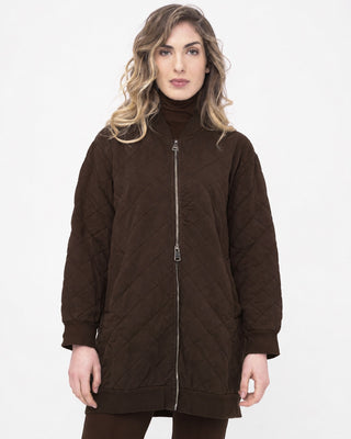 A woman wearing a dark brown quilted zip-front jacket over a matching brown turtleneck, photographed against a plain light gray studio background.
