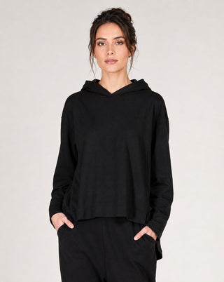 A woman modeling a black long-sleeve cropped hoodie with a relaxed fit, paired with black pants, against a plain light gray studio background.