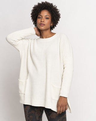 A woman modeling an oversized off-white ribbed knit sweater with front patch pockets, set against a plain light gray studio background.