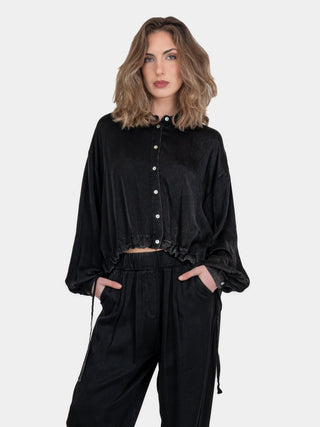 A female model wearing a short bolero shirt with drawstrings and shell buttons, paired with matching pants.