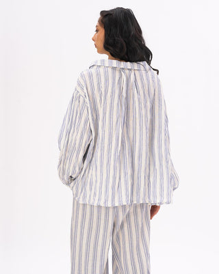 A model wearing a striped crinkled button up peasant shirt with a loose fit The shirt is white with blue stripes and blue pants