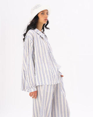 A model wearing a striped crinkled button up peasant shirt with a loose fit The shirt is white with blue stripes and blue pants. she is wearing a hat