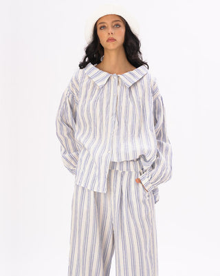 A model wearing a striped crinkled button up peasant shirt with a loose fit and fabricated buttons. The shirt is white with blue stripes and the model is accessorized with a white hat and the same color pants.