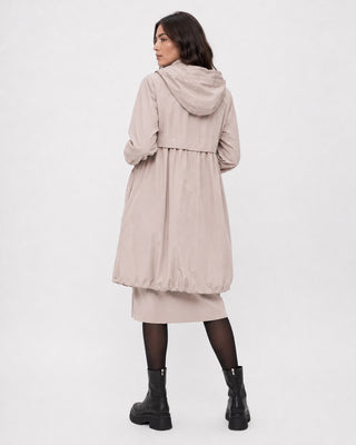 A woman shown from the back wearing a light beige hooded longline coat over a matching midi dress, paired with black tights and black ankle boots, against a plain white studio background.