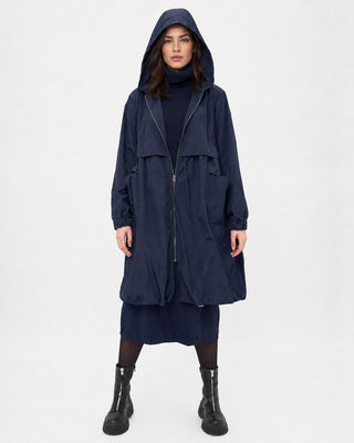 A model wearing a long navy blue hooded windbreaker coat over a matching navy dress, paired with black boots, shot against a plain white studio background.