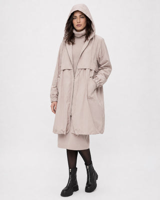 A model wearing a long, light beige hooded coat over a matching beige turtleneck and skirt, paired with black tights and black leather ankle boots, against a plain white studio background.