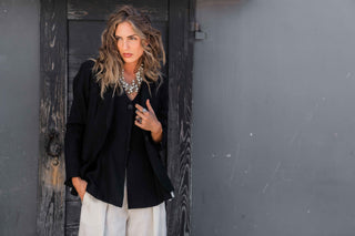 Woman in a black jacket and white pants standing against a dark wall.