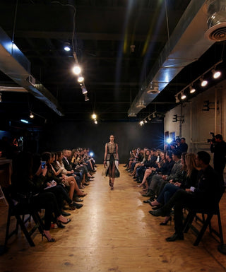 A runway show in a dark industrial loft with a wooden floor, seated audience on both sides, and a model walking down the center aisle wearing a long sheer black dress with a metallic gold patterned bodice.
