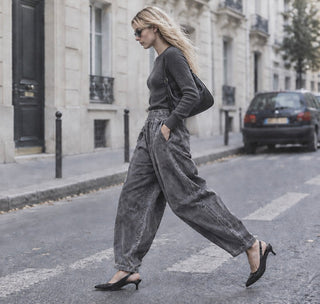 A woman walking across a city street wearing a dark gray long-sleeve top and matching wide-leg high-waisted trousers, paired with black slingback heels and carrying a black shoulder bag.