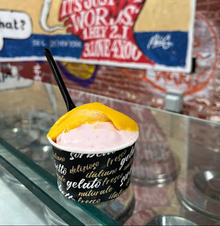 A cup of pink gelato topped with a yellow mango slice and a black straw, set on a glass counter in front of a colorful graffiti-style sign.