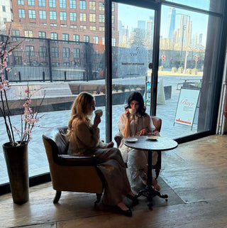 Two women sitting in armchairs by a large window in a modern cafe, drinking coffee from white cups on a small round table. The background shows an urban outdoor plaza and buildings visible through the glass.