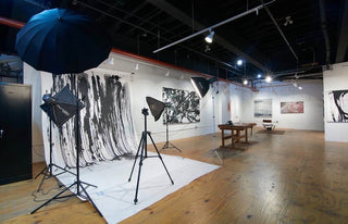 An art gallery or studio interior with photography equipment including softboxes, a tripod, and a large umbrella, alongside framed black-and-white abstract wall art and a wooden table.