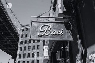 Black and white photo of a sign with 'Baci' on a building with a bridge in the background.