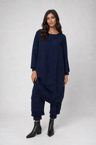 A woman modeling a dark navy blue two-piece outfit consisting of a loose long-sleeve tunic top and matching draped pants, paired with black ankle boots, against a light gray studio background.