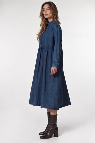 A woman modeling a long-sleeve, high-neck midi dress made of dark blue denim, paired with dark brown leather ankle boots, photographed against a seamless light gray studio background.