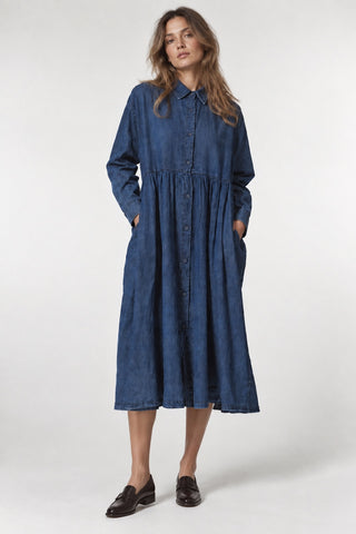 A woman modeling a long-sleeve, button-front midi dress in blue denim or chambray fabric, paired with dark brown loafers, against a plain light gray studio background.