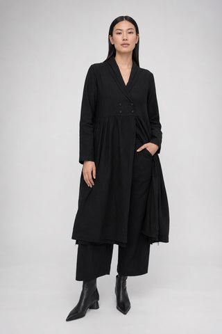 A model wearing a long black double-breasted wrap coat with wide sleeves, paired with black wide-leg trousers and black pointed-toe ankle boots, photographed against a neutral light gray studio background.