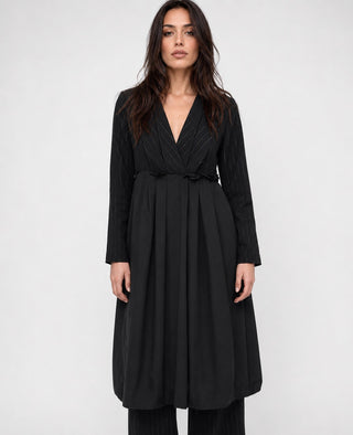 A woman modeling a black long-sleeve wrap dress with a textured pattern on the bodice and sleeves, featuring a tie waist and a flowing skirt, set against a plain light gray background.