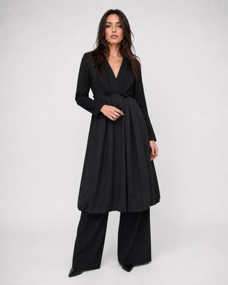 A woman modeling a black long-sleeve wrap dress with a matching tie belt and wide-leg pleated trousers, photographed against a light gray studio background.