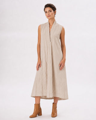 A woman modeling a sleeveless, high-neck, midi-length beige dress with a textured crinkled fabric, paired with tan ankle boots, against a plain white studio background.