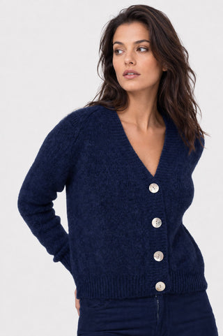 A woman wearing a navy blue knit cardigan sweater with silver buttons, shown from the waist up against a light gray studio background.