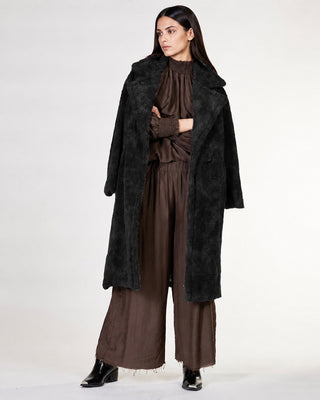 A model stands against a light gray studio background wearing a long black faux-fur coat over a brown pleated top and wide-leg brown pants, with black pointed shoes.