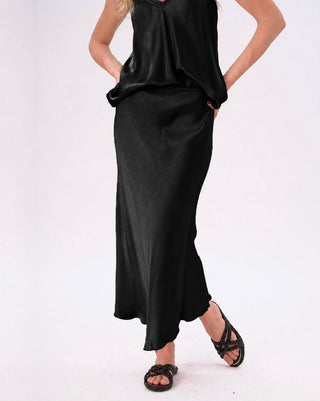 A woman modeling a black satin-like sleeveless top and matching long skirt, paired with black strappy sandals, against a plain white studio background.