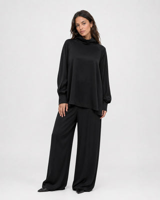 A woman modeling a black long-sleeve cowl-neck top with matching wide-leg pants against a plain light gray studio background.