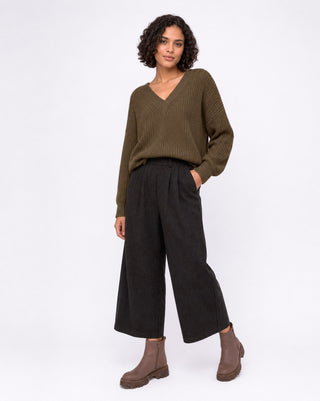 A model wearing an olive green ribbed knit sweater, black wide-leg cropped trousers, and brown ankle boots, standing against a light lavender studio background.