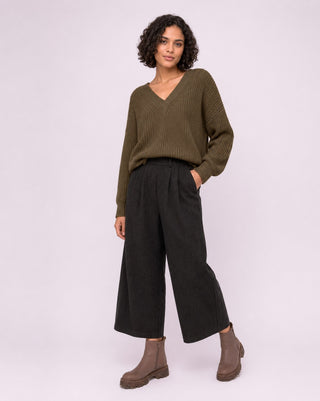 A model wearing an olive green ribbed knit sweater, black wide-leg cropped trousers, and brown ankle boots, standing against a light lavender studio background.