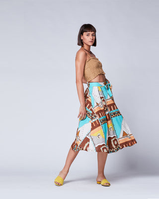 A model wearing a maxi skirt with a belted waistband, featuring a geometric Aztec pattern in multicolor. She is also wearing a tan top and yellow slides.