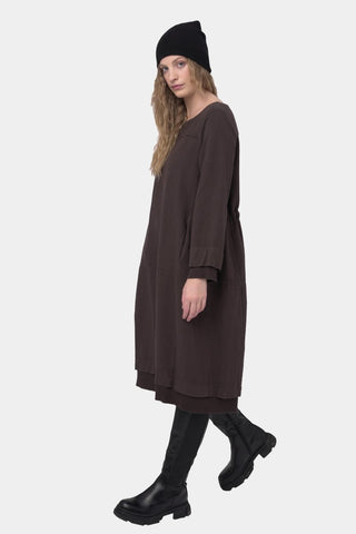 A woman modeling a dark brown two-piece outfit consisting of a loose long-sleeve tunic top, paired with black boots, against a light gray studio background.