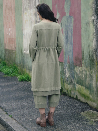 A woman modeling an olive green two-piece outfit consisting of a loose long-sleeve tunic top, paired with brown boots, outdoors. 