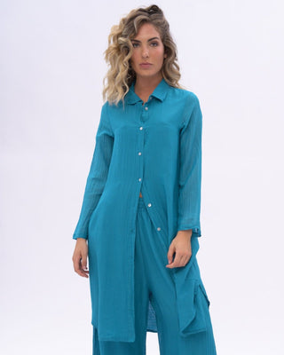 A woman modeling a blue two-piece outfit consisting of a long-sleeve button-front tunic and matching wide-leg pants, photographed against a plain light gray studio background.